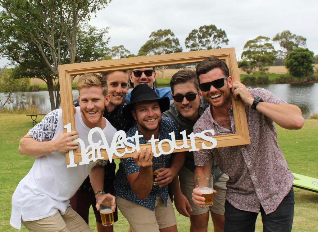 A group of men hold up the Harvest Tours logo with beers